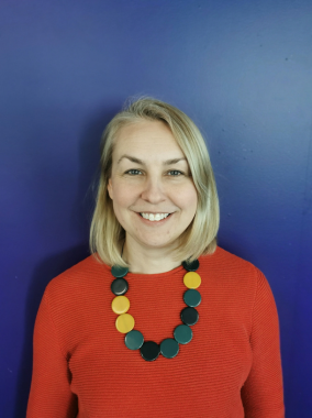 Portrait of a woman wearing a red jumper and a necklace made up of different coloured discs. She is smiling at the camera