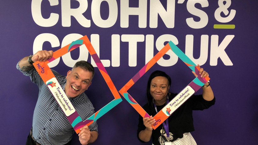 Two people in front of a purple wall with the Crohn's & Colitis UK logo holding VolunteerWeek selfie frame props.